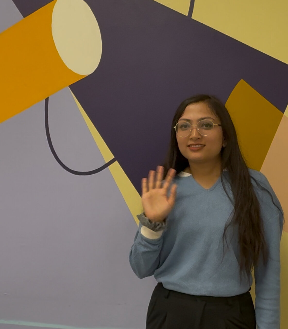 a young women of south east asian decent with glasses waving in front of colourful modern art 